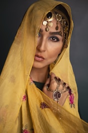 A person adorned with a yellow scarf featuring embroidered floral patterns, partially covering their head and shoulders. They wear ornate jewelry including detailed gold headpieces with dangling coins, and an intricate hand accessory with large stone embellishments. Their makeup includes defined eyes with a hint of blue.