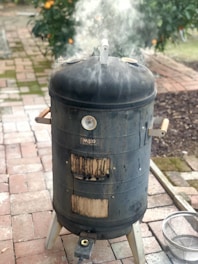 A black vertical smoker is releasing smoke as it stands on a brick patio. It has a temperature gauge and wooden handles on either side. There is vegetation with orange fruit in the background, and a metal bowl sitting on the ground beside the smoker.