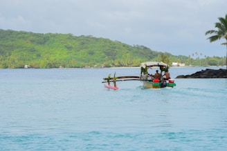 A peaceful tropical scene features a small motorized outrigger boat decorated with palm leaves and flowers, carrying several people in casual attire. The turquoise waters are calm, with a lush green island visible in the background. A few houses are scattered along the shoreline. A palm tree on the right adds to the tropical setting, and the sky is overcast yet bright.