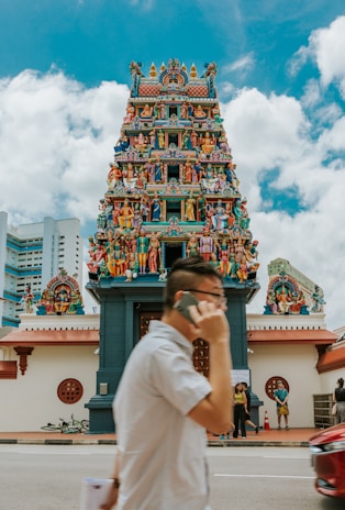 A colorful Hindu temple adorned with intricate carvings and numerous statues of deities and mythological figures is depicted. A person in the foreground is talking on a mobile phone while walking past the temple. The sky is partly cloudy, and there are a few people standing near the entrance of the temple.