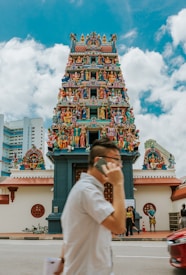 A colorful Hindu temple adorned with intricate carvings and numerous statues of deities and mythological figures is depicted. A person in the foreground is talking on a mobile phone while walking past the temple. The sky is partly cloudy, and there are a few people standing near the entrance of the temple.