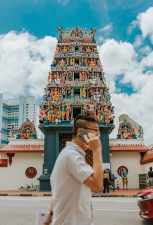 A colorful Hindu temple adorned with intricate carvings and numerous statues of deities and mythological figures is depicted. A person in the foreground is talking on a mobile phone while walking past the temple. The sky is partly cloudy, and there are a few people standing near the entrance of the temple.
