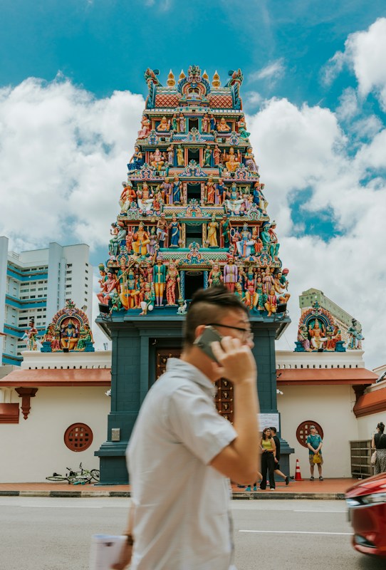 A colorful Hindu temple adorned with intricate carvings and numerous statues of deities and mythological figures is depicted. A person in the foreground is talking on a mobile phone while walking past the temple. The sky is partly cloudy, and there are a few people standing near the entrance of the temple.