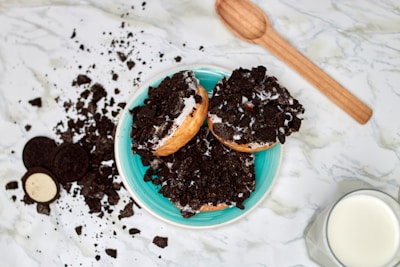 Three donuts are covered in crushed chocolate cookie pieces, placed on a turquoise plate on a marble surface. Beside the plate, some whole and halved chocolate sandwich cookies and a wooden spoon are scattered, alongside a glass of milk.