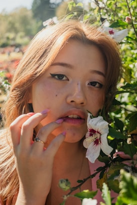 A person with long hair is surrounded by green foliage and white flowers. The sunlight creates a warm glow on their face, highlighting delicate features and making the eyes stand out. A hand with pink nail polish gently touches the lips.