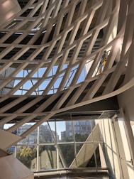 The image captures a modern architectural interior with a series of white, curved structural elements that crisscross diagonally against a backdrop of large windows. Through the windows, an urban cityscape with buildings and greenery is visible under a clear blue sky. The pattern formed by the intersecting structures creates a dynamic and artistic visual effect.