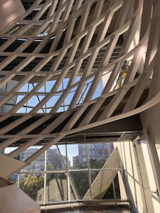 The image captures a modern architectural interior with a series of white, curved structural elements that crisscross diagonally against a backdrop of large windows. Through the windows, an urban cityscape with buildings and greenery is visible under a clear blue sky. The pattern formed by the intersecting structures creates a dynamic and artistic visual effect.
