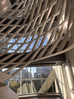 The image captures a modern architectural interior with a series of white, curved structural elements that crisscross diagonally against a backdrop of large windows. Through the windows, an urban cityscape with buildings and greenery is visible under a clear blue sky. The pattern formed by the intersecting structures creates a dynamic and artistic visual effect.