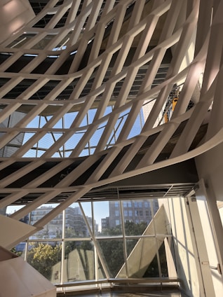 The image captures a modern architectural interior with a series of white, curved structural elements that crisscross diagonally against a backdrop of large windows. Through the windows, an urban cityscape with buildings and greenery is visible under a clear blue sky. The pattern formed by the intersecting structures creates a dynamic and artistic visual effect.