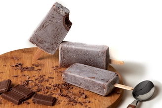 Three chocolate popsicles are arranged on a wooden platter. One popsicle has a bite taken out of it and is leaning against another. Chocolate shavings and pieces of chocolate are scattered on the platter, and a spoon is placed beside them.