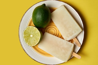 A circular white plate with a brown rim holds two cream-colored popsicles, alongside a whole lime and a lime half. The background is a bright yellow surface, complementing the citrus theme.