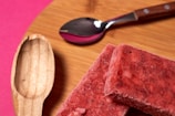 On a wooden surface, two red frozen dessert bars are placed with a metal spoon and a wooden scoop nearby. The background features a vibrant pink color.