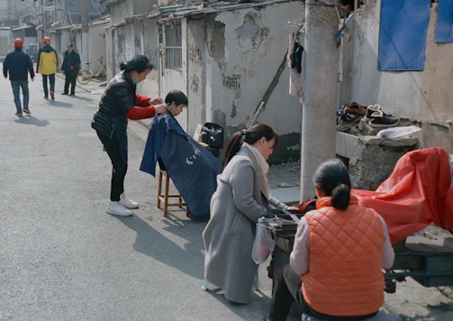 An outdoor barbershop scene with a barber cutting hair for a seated customer on a sidewalk. The setup is simple, with a chair for the customer and minimal equipment. Surrounding them, a few people are present, some walking in the background wearing various jackets and hats. The environment includes old, worn buildings with exposed brick and weathered surfaces. Clothing items are draped on a line or pole nearby.