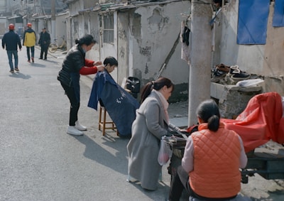An outdoor barbershop scene with a barber cutting hair for a seated customer on a sidewalk. The setup is simple, with a chair for the customer and minimal equipment. Surrounding them, a few people are present, some walking in the background wearing various jackets and hats. The environment includes old, worn buildings with exposed brick and weathered surfaces. Clothing items are draped on a line or pole nearby.