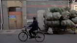 An individual rides a bicycle past a makeshift vehicle loaded with numerous large, bulging bags covered in green tarp. The scene is set against a background of dark brick walls and a closed wooden door with a sign. It appears to be an urban environment, suggesting elements of transportation and manual labor.