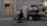 An individual rides a bicycle past a makeshift vehicle loaded with numerous large, bulging bags covered in green tarp. The scene is set against a background of dark brick walls and a closed wooden door with a sign. It appears to be an urban environment, suggesting elements of transportation and manual labor.
