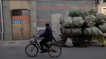 An individual rides a bicycle past a makeshift vehicle loaded with numerous large, bulging bags covered in green tarp. The scene is set against a background of dark brick walls and a closed wooden door with a sign. It appears to be an urban environment, suggesting elements of transportation and manual labor.