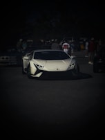 A sleek white sports car is prominently displayed on a dark asphalt surface. Several people stand in the background, some wearing casual clothing and caps, while others appear to be in conversation. The scene suggests a casual outdoor gathering or car show.