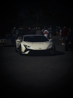 A sleek white sports car is prominently displayed on a dark asphalt surface. Several people stand in the background, some wearing casual clothing and caps, while others appear to be in conversation. The scene suggests a casual outdoor gathering or car show.