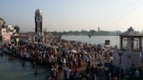 A large number of people gather around a clock tower near a riverbank, engaging in social and religious activities. Some are bathing in the river while others stand or walk along the pier. The scene is lively with many colorful dresses and a mix of traditional and modern attire. Structures and temples are visible in the background, and the presence of greenery can be seen across the river.
