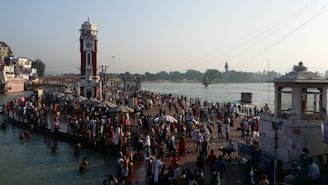 A large number of people gather around a clock tower near a riverbank, engaging in social and religious activities. Some are bathing in the river while others stand or walk along the pier. The scene is lively with many colorful dresses and a mix of traditional and modern attire. Structures and temples are visible in the background, and the presence of greenery can be seen across the river.