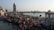 A large number of people gather around a clock tower near a riverbank, engaging in social and religious activities. Some are bathing in the river while others stand or walk along the pier. The scene is lively with many colorful dresses and a mix of traditional and modern attire. Structures and temples are visible in the background, and the presence of greenery can be seen across the river.