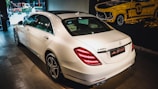 A white luxury sedan is parked indoors, showcasing its sleek body design and distinctive rear lights. The vehicle's branding is visible on the trunk. The setting appears to be a showroom with a stylized mural of a classic car on the wall.
