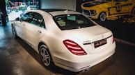 A white luxury sedan is parked indoors, showcasing its sleek body design and distinctive rear lights. The vehicle's branding is visible on the trunk. The setting appears to be a showroom with a stylized mural of a classic car on the wall.