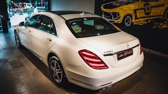A white luxury sedan is parked indoors, showcasing its sleek body design and distinctive rear lights. The vehicle's branding is visible on the trunk. The setting appears to be a showroom with a stylized mural of a classic car on the wall.