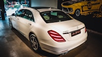 A white luxury sedan is parked indoors, showcasing its sleek body design and distinctive rear lights. The vehicle's branding is visible on the trunk. The setting appears to be a showroom with a stylized mural of a classic car on the wall.