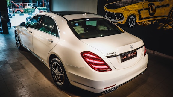 A white luxury sedan is parked indoors, showcasing its sleek body design and distinctive rear lights. The vehicle's branding is visible on the trunk. The setting appears to be a showroom with a stylized mural of a classic car on the wall.
