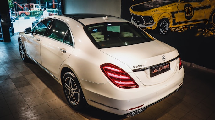 A white luxury sedan is parked indoors, showcasing its sleek body design and distinctive rear lights. The vehicle's branding is visible on the trunk. The setting appears to be a showroom with a stylized mural of a classic car on the wall.