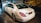 A white luxury sedan is parked indoors, showcasing its sleek body design and distinctive rear lights. The vehicle's branding is visible on the trunk. The setting appears to be a showroom with a stylized mural of a classic car on the wall.