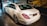 A white luxury sedan is parked indoors, showcasing its sleek body design and distinctive rear lights. The vehicle's branding is visible on the trunk. The setting appears to be a showroom with a stylized mural of a classic car on the wall.
