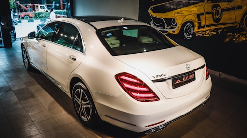 A white luxury sedan is parked indoors, showcasing its sleek body design and distinctive rear lights. The vehicle's branding is visible on the trunk. The setting appears to be a showroom with a stylized mural of a classic car on the wall.