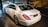 A white luxury sedan is parked indoors, showcasing its sleek body design and distinctive rear lights. The vehicle's branding is visible on the trunk. The setting appears to be a showroom with a stylized mural of a classic car on the wall.