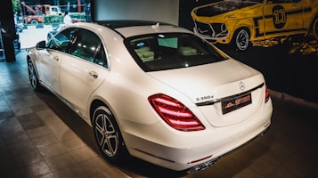 A white luxury sedan is parked indoors, showcasing its sleek body design and distinctive rear lights. The vehicle's branding is visible on the trunk. The setting appears to be a showroom with a stylized mural of a classic car on the wall.