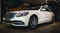 A white luxury sedan is parked indoors, featuring a sleek design with a prominent front grille and stylish alloy wheels. The car is displayed under warm indoor lighting with a mural of a vintage car in the background.