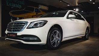 A white luxury sedan is parked indoors, featuring a sleek design with a prominent front grille and stylish alloy wheels. The car is displayed under warm indoor lighting with a mural of a vintage car in the background.