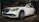 A white luxury sedan is parked indoors, featuring a sleek design with a prominent front grille and stylish alloy wheels. The car is displayed under warm indoor lighting with a mural of a vintage car in the background.