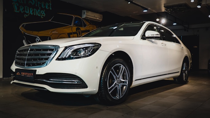 A white luxury sedan is parked indoors, featuring a sleek design with a prominent front grille and stylish alloy wheels. The car is displayed under warm indoor lighting with a mural of a vintage car in the background.