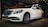 A white luxury sedan is parked indoors, featuring a sleek design with a prominent front grille and stylish alloy wheels. The car is displayed under warm indoor lighting with a mural of a vintage car in the background.
