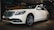 A white luxury sedan is parked indoors, featuring a sleek design with a prominent front grille and stylish alloy wheels. The car is displayed under warm indoor lighting with a mural of a vintage car in the background.