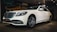 A white luxury sedan is parked indoors, featuring a sleek design with a prominent front grille and stylish alloy wheels. The car is displayed under warm indoor lighting with a mural of a vintage car in the background.