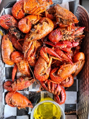 A platter filled with seasoned and cooked crawfish, displaying a vibrant red-orange color. The crawfish are presented on a checkered black and white paper, accompanied by a small clear dish containing melted butter or oil.