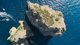An aerial view of two large rocky islands surrounded by deep blue water. The islands are covered with patches of green vegetation. Several small boats are navigating through the water, creating white trails on the surface.