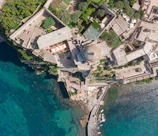 An aerial view of a coastal region featuring a complex of large buildings with beige and reddish roofs surrounded by lush green trees. The architecture is situated atop a rocky coastline adjacent to clear blue-green water. A small dock with boats is visible at the edge of the water.