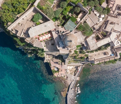 An aerial view of a coastal region featuring a complex of large buildings with beige and reddish roofs surrounded by lush green trees. The architecture is situated atop a rocky coastline adjacent to clear blue-green water. A small dock with boats is visible at the edge of the water.