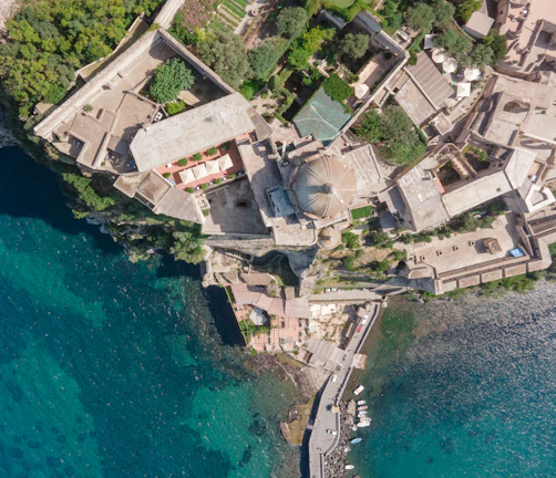 An aerial view of a coastal region featuring a complex of large buildings with beige and reddish roofs surrounded by lush green trees. The architecture is situated atop a rocky coastline adjacent to clear blue-green water. A small dock with boats is visible at the edge of the water.