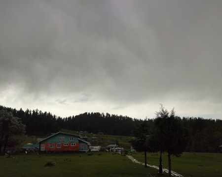 A serene countryside scene with a large, overcast sky dominating the view. A distinctive green-and-orange house is situated in the foreground, surrounded by lush greenery and a few trees. In the background, a dense forest outlines the horizon.